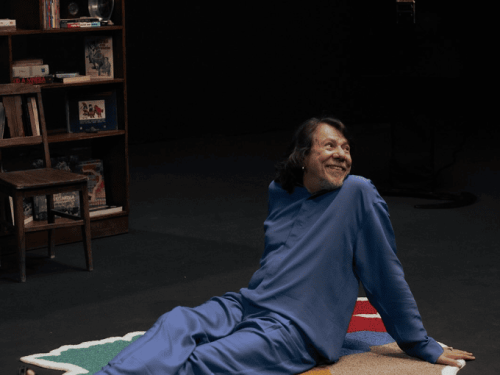 O cantor Lenine aparece sentado e relaxado sobre um tapete colorido e recortado em formato abstrato, no centro de um cenário escuro. Ele veste um conjunto azul vibrante e olha para o lado com um sorriso aberto. Ao fundo, à esquerda, há uma estante de madeira com livros e objetos decorativos, como uma caveira colorida e um prato metálico, criando uma atmosfera íntima e artística, como se ele estivesse em seu próprio estúdio ou biblioteca pessoal.