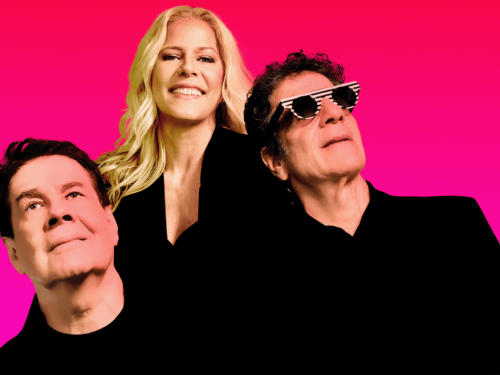 A imagem apresenta os três integrantes da banda **Kid Abelha** — Paula Toller, George Israel e Bruno Fortunato — posando contra um fundo rosa vibrante. **Paula Toller** está ao centro, sorrindo e com seus longos cabelos loiros em destaque. À sua esquerda, **George Israel** usa óculos escuros com armação listrada e olha para cima, enquanto **Bruno Fortunato** aparece à direita, também olhando para o alto. Todos vestem roupas pretas, criando um contraste moderno e minimalista com o fundo colorido.