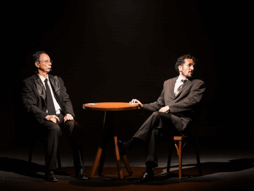 Dois homens vestidos com ternos escuros e gravatas estão sentados em volta de uma pequena mesa redonda de madeira. O palco está escuro, com um foco de luz quente centralizado sobre eles. O homem à esquerda mantém uma expressão séria e pensativa, enquanto o da direita está com as pernas cruzadas e parece falar ou reagir a algo, criando um clima de diálogo teatral.