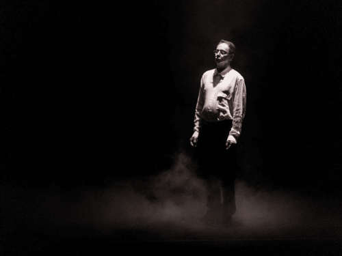  Imagem em preto e branco de um homem de óculos e camisa clara, em pé no centro de um palco escuro. Ele está sob um foco de luz vertical, cercado por uma fumaça densa que cobre o chão, criando uma atmosfera dramática e melancólica.