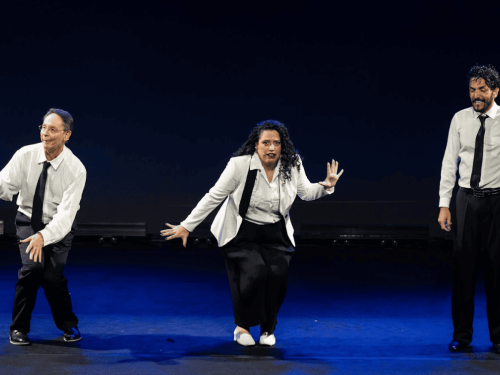 Três artistas — dois homens e uma mulher — realizam uma performance em um palco com fundo escuro e iluminação azulada. Todos vestem calças pretas e camisas brancas com gravatas. Eles exibem expressões dinâmicas e poses coreografadas, sugerindo um momento de teatro, dança ou improviso.