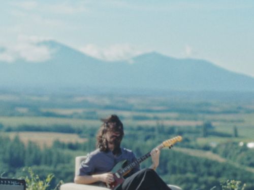 Fotografia do cantor Mateus Asato sentado em um sofá com paisagem de fundo