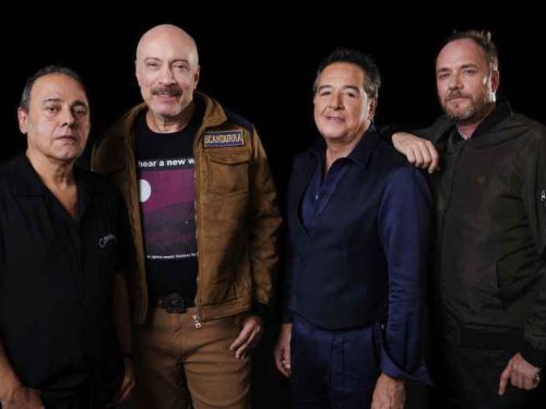 A fotografia promocional do grupo Ira! destaca os quatro integrantes principais da banda de rock posicionados lado a lado, em meio corpo, contra um fundo inteiramente preto. A iluminação direcionada foca nos músicos, criando um contraste nítido com a escuridão ao redor e ressaltando suas expressões e vestimentas. No extremo esquerdo, o primeiro integrante veste uma camisa de botão preta de mangas curtas, mantendo uma expressão séria e os braços estendidos ao longo do corpo. Ao seu lado, o segundo músico ex