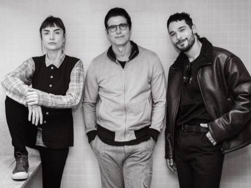 Fotografia em preto e branco de três pessoas posando lado a lado em frente a uma parede de azulejos claros. À esquerda, a atriz Maria Casadevall usa colete escuro sobre camisa xadrez e apoia um dos pés em uma bancada. No centro, o ator Reynaldo Gianecchini sorri de óculos, vestindo jaqueta de zíper e calça clara. À direita, um homem jovem de barba sorri de jaqueta de couro e roupa preta.