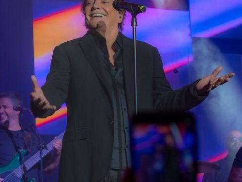 Fotografia em plano médio do cantor Fábio Jr. sorrindo no palco durante um show. No centro, ele veste terno e camisa pretos, com os braços abertos em um gesto acolhedor diante do microfone. O fundo traz painéis de LED coloridos com faixas em tons de azul, rosa e amarelo, além de fumaça cenográfica. À esquerda, na penumbra, aparece um músico tocando contrabaixo elétrico verde e, em primeiro plano, vê-se o topo desfocado de celulares do público registrando a apresentação.