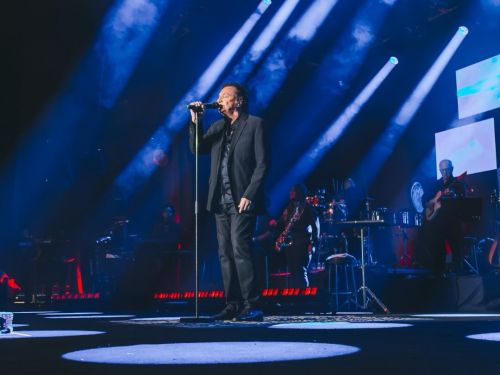 Fotografia em plano geral e contra-plongê do cantor Fábio Jr. se apresentando no palco durante um show. No centro, o artista veste terno e camisa pretos, segurando o microfone no pedestal enquanto canta de perfil. O palco é iluminado por fortes feixes de luz azul que cortam o cenário de cima para baixo, criando círculos luminosos no chão escuro. Ao fundo, na penumbra, é possível ver os músicos da banda com seus instrumentos, como saxofone e guitarra, além de telas de LED integradas à cenografia.