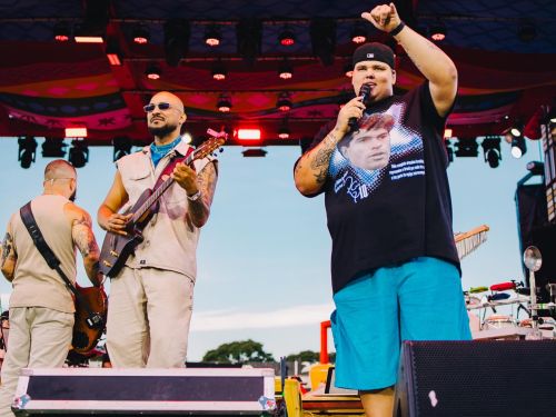 Fotografia em plano médio de integrantes do grupo Menos é Mais no palco durante um show. À direita, o vocalista canta com o microfone na mão e o braço erguido, vestindo boné, camiseta preta com a estampa de Maradona e bermuda azul-turquesa. À esquerda, outro integrante de óculos escuros e roupa bege sorri enquanto toca violão, acompanhado por um baixista de costas ao fundo. O palco traz caixas de som em primeiro plano e iluminação vermelha na cobertura.