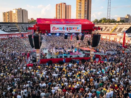 Fotografia aérea em plano geral mostrando uma multidão lotando o gramado de um estádio de futebol durante o evento musical "Churrasquinho". No centro, destaca-se um grande palco coberto por uma lona vermelha, decorado com uma faixa colorida onde se lê o nome do evento. Artistas se apresentam no palco circular, rodeados por caixas de som suspensas. Ao redor do palco, milhares de pessoas aparecem aglomeradas sob a luz do dia, muitas vestindo chapéus amarelos.