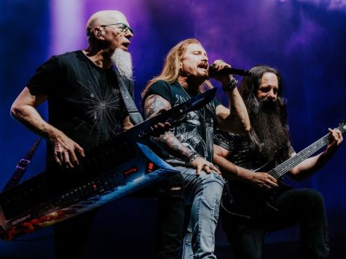 Três integrantes do Dream Theater em ação no palco sob luzes roxas. À esquerda, Jordan Rudess toca seu teclado personalizado (keytar); ao centro, o vocalista James LaBrie canta com intensidade no microfone; e à direita, John Petrucci toca sua guitarra. O clima é de pura energia e concentração musical.