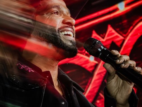 Foto em plano médio e ângulo contra-plongée do cantor Alexandre Pires sorrindo e cantando em um microfone preto. Ele veste jaqueta escura com detalhes bordados e um chapéu preto. O fundo traz painéis de LED com iluminação vermelha intensa e um efeito dinâmico de rastro de movimento desfocado na lateral esquerda.