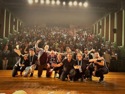 Fotografia panorâmica tirada a partir do palco de um teatro com piso de madeira. Em primeiro plano, um grupo de cerca de dez pessoas — incluindo os atores Reynaldo Gianecchini e Maria Casadevall — sorri e posa para a foto. Ao fundo, a plateia lotada ocupa as poltronas em uma arquibancada inclinada, com muitas pessoas acenando e segurando celulares com as luzes das lanternas acesas, criando vários pontos brilhantes na penumbra do auditório.