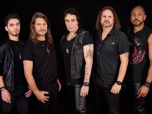 A fotografia promocional da banda de metal Angra retrata os cinco integrantes do grupo posicionados em pé, lado a lado, em plano médio e contra um fundo inteiramente preto. A iluminação clara e difusa incide diretamente sobre os músicos, destacando os detalhes de suas expressões e vestimentas, compostas predominantemente por peças de roupas pretas de estilo casual e rockeiro. No centro da composição, posiciona-se um dos integrantes com olhar fixo na câmera, exibindo cabelos escuros repicados na altura dos 