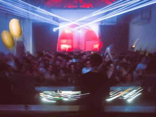 Foto desfocada e com movimento capturando a energia de uma festa em um local industrial. No primeiro plano, o equipamento do DJ exibe rastros de luz branca e colorida. À esquerda, dois balões amarelos flutuam sobre uma multidão densa de pessoas com os braços levantados. Ao fundo, luzes de laser azuis e brancas cruzam o espaço, iluminando estruturas arqueadas com luz vermelha intensa.
