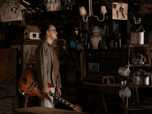 Fotografia em ambiente rústico com iluminação dramática. No centro, de perfil, um homem sorridente segura uma guitarra semiacústica de cor sunburst (laranja e marrom). O cenário ao fundo é escuro e repleto de elementos vintage, como discos de vinil pendurados no teto (incluindo um do grupo 14 Bis), um piano antigo, bules de metal e objetos de madeira, criando uma atmosfera nostálgica e artística.