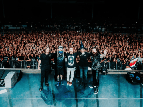 A banda completa está posicionada na beira do palco, de frente para uma multidão imensa que lota a arena. Os cinco músicos estão abraçados, sorrindo e celebrando o fim do show, enquanto o público ao fundo aparece com os braços levantados. Jordan Rudess destaca-se usando um chapéu de mago azul.