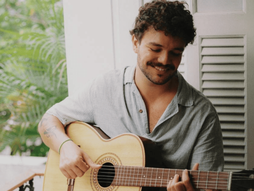 A imagem, com tons suaves e naturais, mostra o músico sorrindo levemente enquanto olha para as cordas de um violão acústico. Ele veste uma camisa listrada de manga curta e está posicionado em frente a uma janela com venezianas brancas abertas. Ao fundo, à esquerda, é possível ver a folhagem verde de palmeiras, criando uma ambientação tranquila e iluminada pela luz do dia.