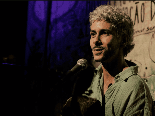 A imagem mostra o artista de perfil parcial, sorrindo suavemente enquanto canta em um microfone e toca violão. Ele tem cabelos curtos, cacheados e descoloridos, e veste uma camisa de tecido leve na cor verde-água. O fundo é escuro, com uma iluminação dramática que destaca tons de roxo à esquerda e uma parede com pichações e frases artísticas à direita, criando uma estética urbana e acolhedora de show ao vivo.