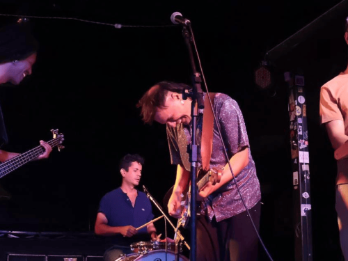 A imagem captura quatro músicos em um palco com iluminação focada e fundo escuro. No centro, o guitarrista — com uma camisa brilhante prateada — aparece inclinado sobre seu instrumento em um momento de entrega. À esquerda, um baixista sorri enquanto toca; ao fundo, o baterista mantém o ritmo concentrado; e à direita, o tecladista acompanha a performance com uma partitura à frente. O clima é de descontração e sintonia entre os artistas durante o show.