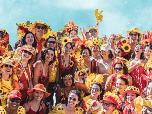 A imagem mostra um grande grupo de foliões sorridentes, a maioria mulheres, celebrando o Carnaval. Eles vestem fantasias vibrantes nas cores vermelho e amarelo, com destaque para diversos adereços de girassóis em chapéus, tiaras e roupas. A atmosfera é de alegria e união, com várias pessoas acenando para a câmera sob um céu azul claro ao fundo. É uma representação típica da energia festiva dos blocos de rua mineiros.