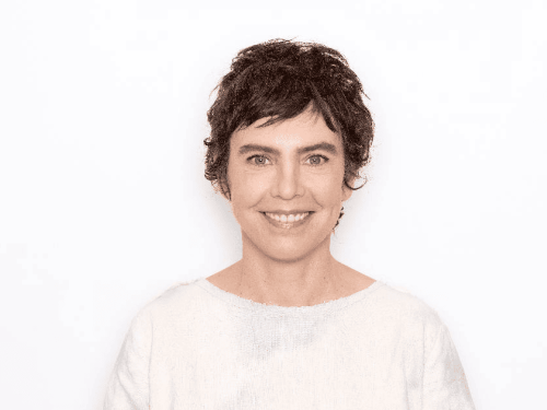 A imagem apresenta um plano médio centralizado da artista sorrindo de forma leve e acolhedora, contra um fundo totalmente branco e infinito. Ela possui cabelos escuros, curtos e levemente repicados, e veste uma blusa branca de tecido simples e textura natural. A iluminação é suave e frontal, destacando seus traços com clareza e transmitindo uma sensação de serenidade e minimalismo.