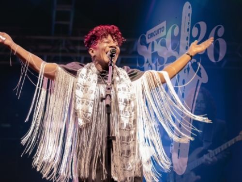 O cartaz apresenta uma cantora negra no centro da imagem, em um momento de grande expressividade durante uma apresentação musical ao vivo. Ela está de olhos fechados e com um largo sorriso, com o microfone posicionado à sua frente e os braços abertos para os lados, transmitindo uma sensação de entrega e conexão com o público. Seu cabelo é curto e está tingido em um tom vibrante de rosa ou vinho. Ela veste uma roupa escura complementada por uma echarpe estampada com padrões que lembram teclas de piano e uma 