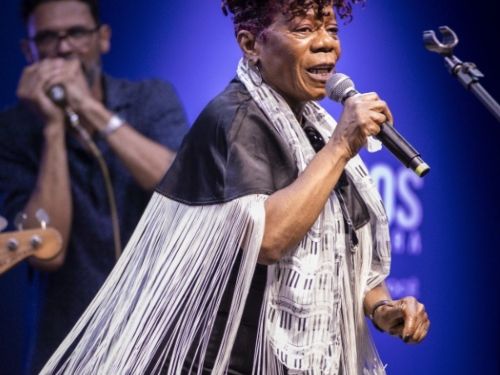 A imagem traz o registro da cantora no mesmo show da primeira foto, com foco em uma expressão dinâmica e enérgica. Ela aparece de perfil para três quartos, ligeiramente inclinada para a frente, segurando o microfone com a mão direita e com a boca aberta, capturando um instante em que canta com intensidade. Seu cabelo curto e cacheado em tom de rosa escuro está bem destacado sob a iluminação do palco. Ela veste o mesmo figurino composto pelo vestido preto, a echarpe com estampa de teclas de piano e a capa d