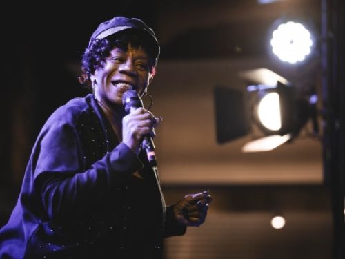 A imagem retrata uma cantora negra sorridente durante uma apresentação musical em um palco intimista. Ela está posicionada do lado esquerdo do enquadramento, voltada levemente em direção ao público, enquanto segura um microfone com a mão direita próximo ao rosto. A artista veste uma roupa preta de mangas longas, detalhada com pequenos pontos brilhantes na lateral, e usa grandes brincos de argola. Na cabeça, ela utiliza uma boina escura com uma faixa brilhante na aba, que deixa à mostra alguns cachos escuro