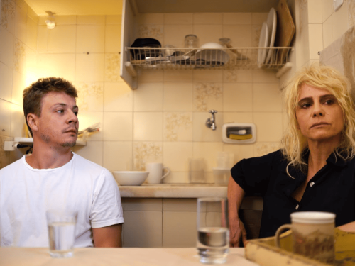 Fotografia em plano médio de um homem e uma mulher sentados lado a lado em uma cozinha de azulejos claros e luz amarelada. À esquerda, o homem jovem de camiseta branca olha de perfil em direção à mulher. À direita, ela tem cabelos loiros desgrenhados, veste uma camisa preta e encara o horizonte com uma expressão séria e cansada. Sobre a mesa em primeiro plano, há copos com água e uma caneca, enquanto ao fundo vê-se uma pia com louças organizadas.