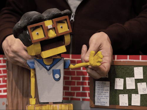 Fotografia em plano detalhe mostrando um boneco de teatro de animação articulado, feito de madeira e papelão. O boneco tem pele amarela, cabelo preto esculpido, óculos de aro quadrado laranja e veste um uniforme escolar cinza e azul. Ao fundo, mãos de um manipulador seguram o boneco e movem um de seus braços. O cenário ao fundo simula uma sala de aula, com uma parede de tijolinhos vermelhos e um pequeno mural verde com papéis em miniatura fixados, incluindo uma lista manuscrita intitulada "Combinados".