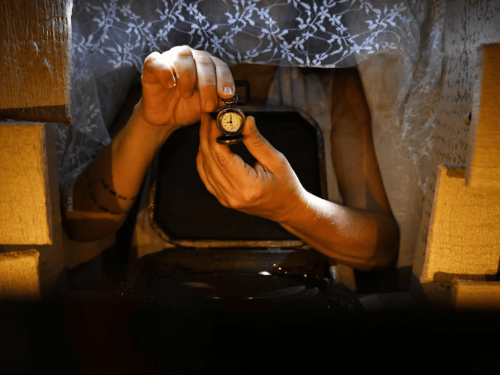Fotografia em plano detalhe e iluminação cênica quente focada nas mãos de uma pessoa. Ela segura e abre um pequeno relógio de bolso antigo. As mãos saem de dentro de uma mala escura aberta. Acima, há uma cortina de renda branca com padrões florais que cobre o rosto da pessoa. As laterais da cena são emolduradas por blocos e paredes de textura rústica em tons de bege, criando a sensação de um minicenário ou caixa de teatro.