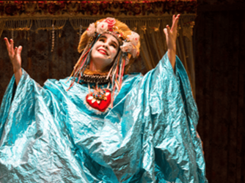 Imagem horizontal mostra uma atriz no palco, do busto para cima, caracterizada com um figurino teatral exuberante. Ela é uma mulher branca, com o rosto pintado de branco e bochechas rosadas, que sorri olhando para o alto com os braços abertos e as mãos erguidas. Veste uma túnica larga e brilhante em tom azul-celeste, um colar com um grande coração vermelho decorado no centro do peito e um adereço de cabeça dourado adornado com flores e fitas coloridas pendentes.