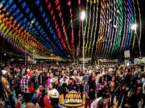 Esta imagem mostra o evento "Arraiá do Granada" acontecendo na prática, registrando uma grande noite festiva com o local completamente lotado de pessoas. O céu noturno está totalmente coberto por fileiras serradas de bandeirinhas juninas multicoloridas que criam um efeito visual vibrante e iluminado sob os refletores. Abaixo delas, o público aparece disperso pela pista, com muitas pessoas vestindo trajes típicos caipiras, como camisas xadrez e chapéus de palha. Centralizado na parte inferior da foto, há um 