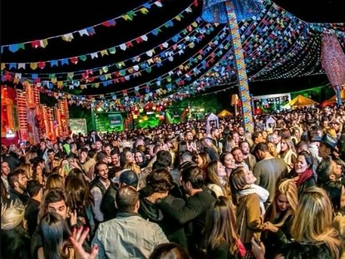 Esta imagem registra a festa "Junina Chevals" acontecendo na prática, mostrando um grande espaço ao ar livre completamente lotado por um público jovem e animado durante a noite. O céu escuro é decorado por extensas fileiras de bandeirinhas juninas coloridas que convergem para uma grande estrutura central iluminada, que se assemelha a um enorme chapéu ou luminária de palha estilizada em tons azuis. Abaixo da decoração, as pessoas aparecem aglomeradas, conversando e dançando, a maioria vestindo roupas de frio
