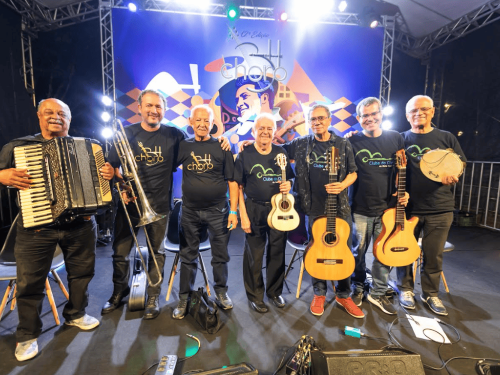 Fotografia colorida de sete músicos homens, adultos e idosos, posando sorridentes lado a lado no palco iluminado da 12ª Edição do BH Choro. Eles vestem calças e camisetas pretas do evento e seguram, da esquerda para a direita: um acordeon, um trombone de vara, um cavaquinho, dois violões e um pandeiro (o terceiro integrante está sem instrumento). Ao fundo, destaca-se um grande painel azul com a ilustração estilizada do músico Pixinguinha tocando flauta.