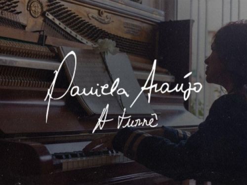 Cartaz horizontal em tons intimistas e rústicos. No lado direito, uma mulher de cabelos longos aparece de perfil, com os braços estendidos tocando as teclas de um piano vertical de madeira escura com o tampo superior aberto, revelando suas cordas e martelos internos. No centro, sobre uma partitura aberta adornada por uma flor branca em cima do piano, destaca-se o nome da artista "Daniela Araújo" escrito em uma caligrafia cursiva branca manuscrita, seguido pelo subtítulo "A Turnê" logo abaixo na mesma tipogr