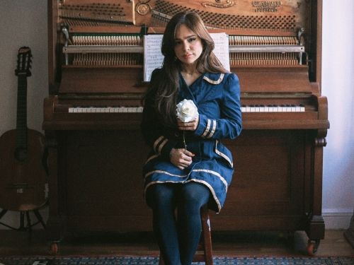 Imagem de Daniela Araújo sentada frente a um piano com uma rosa em sua mão esquerda.