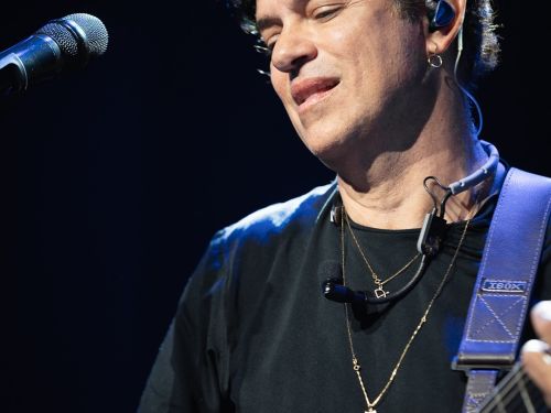 Fotografia em plano médio do cantor Jorge Vercillo tocando violão no palco sob um fundo escuro. Ele está de perfil três quartos, com os olhos semicerrados olhando para o instrumento, veste uma camiseta preta de mangas longas e usa correntes douradas finas com pingentes no pescoço. No canto esquerdo superior, aparece um microfone de palco em destaque, e o cantor utiliza um fone de retorno azul no ouvido esquerdo.
