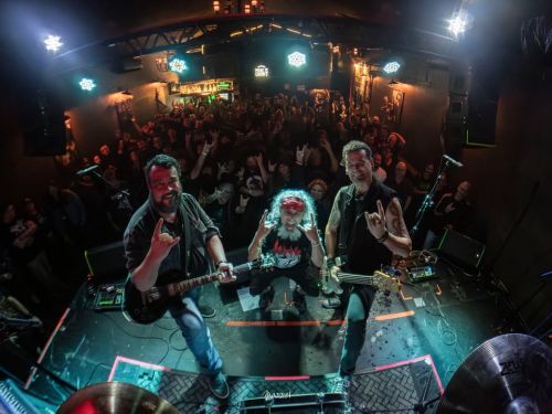 Uma foto tirada de cima para baixo em uma casa de shows escura, com uma lente olho de peixe que distorce as bordas. Em primeiro plano, no palco, três músicos sorridentes estão agachados e fazendo o sinal dos "chifres do rock" com as mãos para a câmera: um guitarrista à esquerda, o vocalista com bandana vermelha ao centro e um baixista à direita. Atrás deles, uma multidão lotada levanta os braços, correspondendo ao gesto. Luzes de palco coloridas iluminam a cena e um banner no bar ao fundo diz "HAKAI CLUB".