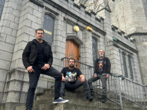 Fotografia colorida tirada de baixo para cima, mostrando três homens com roupas escuras e estilo casual/rocker posando nas escadarias de uma antiga construção de pedras cinzas, que lembra uma igreja gótica. Um homem está de pé à esquerda com a mão na cintura, outro está sentado ao centro dos degraus, e o terceiro está de pé à direita, perto de um corrimão de metal. Ao fundo, destacam-se uma porta de madeira em arco e janelas altas com vitrais.