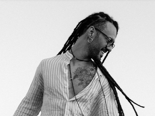 Fotografia em preto e branco com fundo claro. Um homem jovem, com pele negra clara, sorri de perfil olhando para baixo e para a direita. Ele tem cabelos longos com dreadlocks que caem pelas costas e ombros, usa óculos de grau, brinco de argola e um colar discreto. Veste uma camisa clara com listras verticais finas, que está entreaberta, revelando uma tatuagem de girassóis no peito. No canto inferior esquerdo, aparece uma pequena parte do braço de um violão.