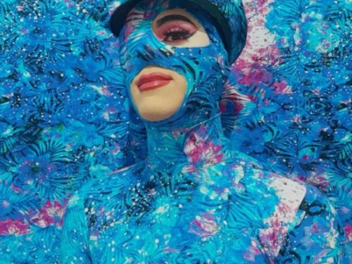 foto de uma mulher drag com fundo azul cumprindo parcialmente seu rosto