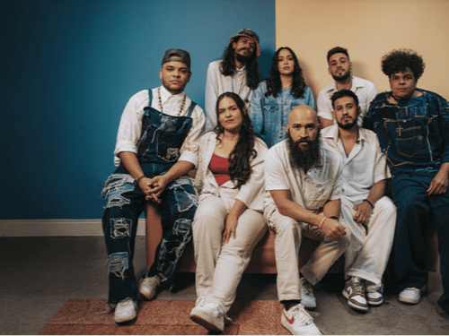 Foto em estúdio de um grupo de oito pessoas vestindo roupas jeans e brancas, posando juntas diante de uma parede dividida entre as cores azul e bege.