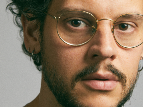 Fotografia de Vinícius de Souza, um homem branco, jovem, usa óculos de aros redondos e finos, bigodes, barba e cabelo castanhos..
