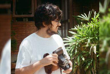 Homem de pele clara, cabelos e barba escuros, usando óculos, segura uma câmera fotográfica antiga.