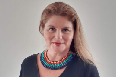 A foto mostra Beatriz, uma mulher branca de cabelos claros. Ela usa uma blusa azul marinho e colar colorido de contas.