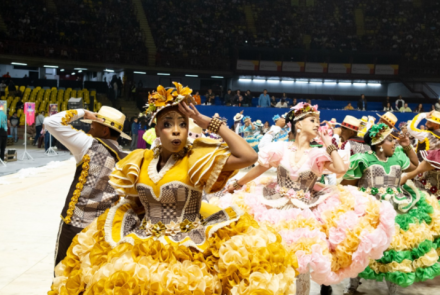 Um grupo de quadrilha junina se apresentando em um tablado, com destaque para uma mulher de vestido amarelo a frente da foto.
