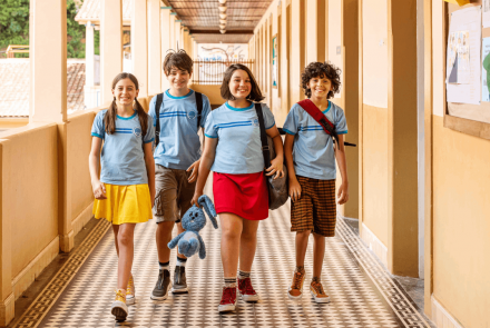 Foto com 4 crianças uniformizadas, caminhando pelo corredor da escola, Magali com camisa de uniforme e saia amarela, Cebolinha ao lado, com camisa de uniforme da escola e bermuda marrom. Mônica com o coelhinho de pelúcia azul, o Sansão, uniformizada, com saia vermelha. Cascão também uniformizado, com bermuda flanelada e xadreza em tons de marrom.