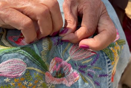 A imagem mostra duas mãos fazendo um bordado de rosas em um tecido azul 