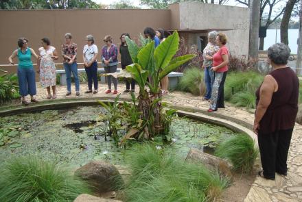 Grupo de pessoas observam um jardim. Na imagem as pessoas estão posicionadas ao redor de um pequeno lago no jardim da MCK. É possível notar plantas verdes ao redor.