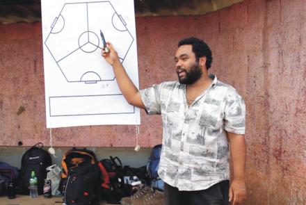 Um homem negro de barba, camisa branca está de pé. Ele aponta para um quadro branco com uma caneta na mão. No quadro branco há um desenho.